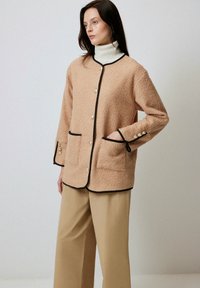 Giacca beige testurizzata con rifiniture nere, chiusure a bottone e due tasche frontali. Abbinata a pantaloni a gamba larga di colore chiaro.