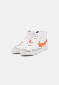 Valkoiset korkeavartiset tennarit, joissa on oranssi Nike-logotikkaus, harmaa mokkanahkainen kärki ja valkoinen kumipohja. Ominaisuuksiin kuuluvat litteät nauhat ja tikatut yksityiskohdat.