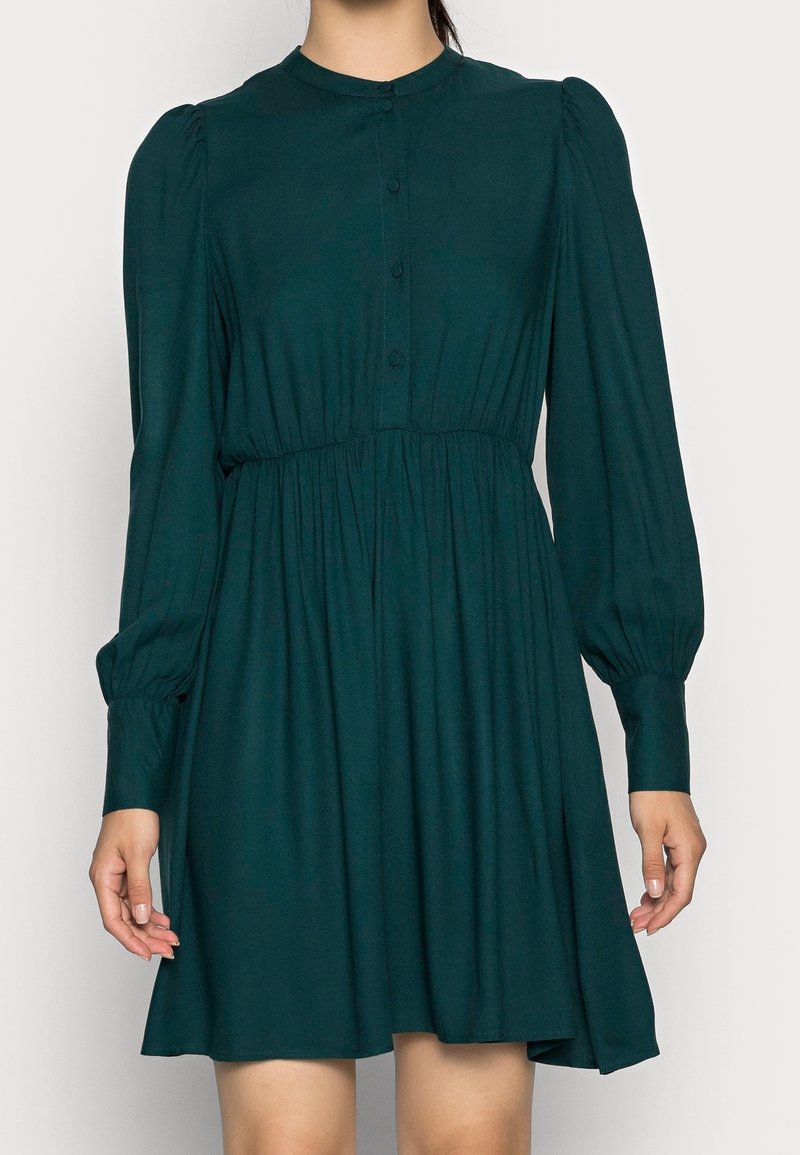 Femme portant une robe vert foncé, à manches longues, boutonnée, longueur genou, avec une taille froncée et des épaules bouffantes.