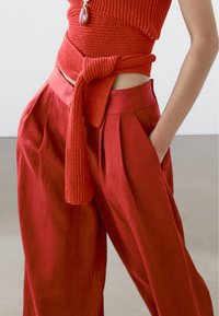 Röd stickad topp med knyt i midjan, tillsammans med lösa röda vida byxor med veck och sidofickor, allt i ett smidigt tyg.