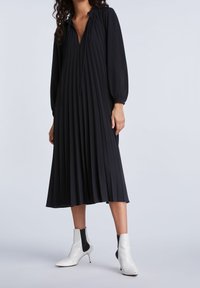 Robe noire plissée avec un décolleté en V, des manches longues et un cordon de serrage. Associée à des bottines blanches à la cheville avec des accents élastiques noirs.