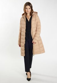 Beige puffjakke med hætte, der har en quiltet design, frontlynlås og sidelommer, lagdelt over et sort outfit og hæle.