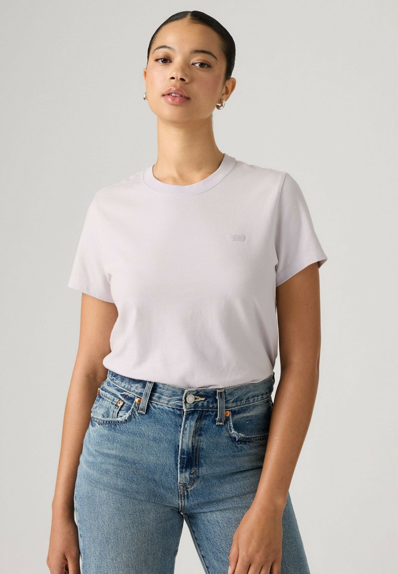 Femme aux cheveux attachés, portant un t-shirt uni lavande clair et un jean bleu taille haute sur un fond neutre.