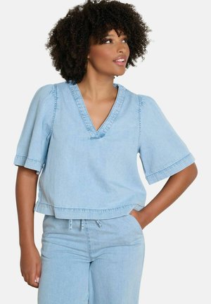 Femme aux cheveux bouclés portant un haut à manches courtes en denim bleu clair avec col en V et un pantalon taille haute assorti, regardant vers sa droite.