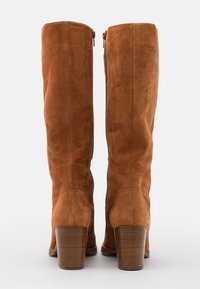 Botas marrones de ante hasta la rodilla con una textura suave, cremallera lateral y tacones de madera gruesa. Diseño sencillo y sin patrones visibles.