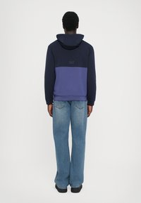 Sweatshirt azul-marinho e roxo com capuz, apresentando um detalhe de logotipo nas costas. Combinado com jeans de perna larga de um azul claro. Textura suave.