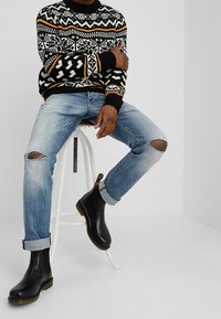 Maglione lavorato a maglia con motivo in bianco e nero, jeans strappati blu chiaro e stivali neri alla caviglia con suole color tan, seduto su uno sgabello bianco.
