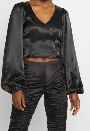 Persona che indossa un crop top in raso nero a maniche lunghe con maniche a sbuffo e pantaloni neri in raso arricciati.