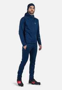 Navy urheilu takki hupulla, vetoketjulliset taskut ja yhteen sopivat istuvat housut. Mallilla on mustat urheilu kengät punaisilla yksityiskohdilla.