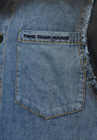 Ενδυματικό τζιν με μπροστινή τσέπη που φέρει μαύρη κεντημένη επιγραφή "Feng CHen WANG" και κίτρινο γαζί, με φθαρμένη άκρη στη δεξιά πλευρά.