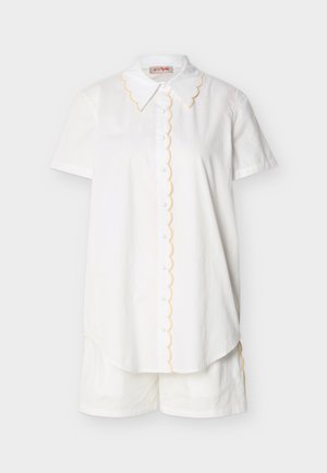 Camisa blanca de manga corta con botones y bordes ondulados, acompañada de pantalones cortos blancos a juego sobre un fondo liso.