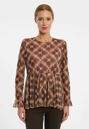 Frau trägt eine langärmelige rosa Bluse mit braunem abstraktem Blumenmuster und Rüschenmanschetten, kombiniert mit dunkler Hose, steht vor weißem Hintergrund.
