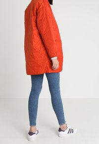 Pomarańczowa kurtka pikowana z długimi rękawami, kontrastowymi mankietami i luźnym krojem, w zestawieniu z niebieskimi skinny jeansami oraz białymi trampkami z fioletowymi akcentami.