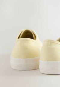 Baskets en toile jaunes avec une surface texturée, semelles en caoutchouc blanches et un design minimaliste. Présente une double couture au niveau du talon.