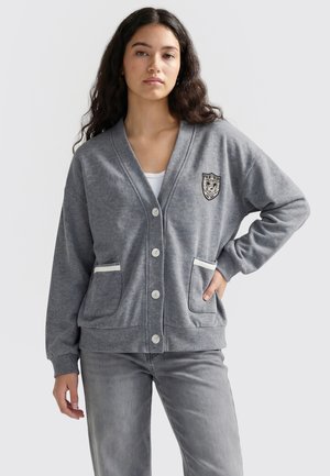 Ung kvinde med langt mørkt hår iført en grå knappet cardigan med frontlommer og emblem, kombineret med grå jeans.