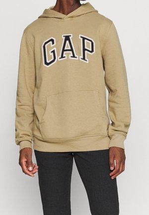 Hombre vistiendo una sudadera con capucha beige de GAP y pantalones negros, de pie contra un fondo blanco liso.