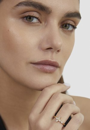 Frau mit reiner Haut und grünen Augen trägt einen goldenen Ring mit vier diamantenförmigen Steinen, die Hand nahe am Kinn vor weißem Hintergrund ruhend.