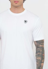 T-shirt bianco a maniche corte realizzato in tessuto liscio, con un piccolo logo a scudo bianco e nero sul lato sinistro del petto.