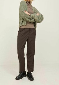 Groene gebreide cardigan met brede mouwen over een bruine coltrui, gecombineerd met donkere bruine tapered broek en zwarte veterschoenen.