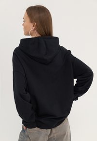 Black hoodie with a large hood, loose fit, and ribbed cuffs. Worn with high-waisted beige pants, showcasing a simple, modern design.
