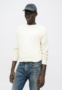 Κρεμ πουλόβερ με ριμπ ύφανση και μακριά μανίκια, συνδυασμένο με μπλε τζιν. Το μοντέλο φοράει σκουρόχρωμο γκρι καπέλο με κεντημένη λεπτομέρεια.