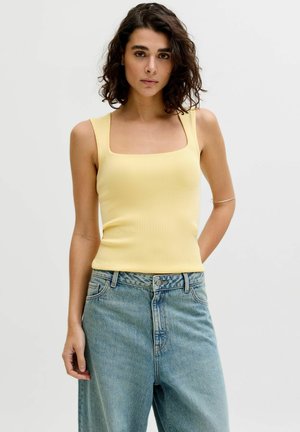 Göndör sötét hajú nő, aki sárga ujjatlan felsőt és világoskék magasderekú farmert visel, egyszínű világos háttér előtt.