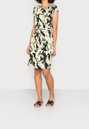 Kobieta stoi ubrana w czarną sukienkę do kolan z zielonym i białym liściastym wzorem, związaną paskiem oraz czarne sandały na płaskim obcasie na jednolitym tle.