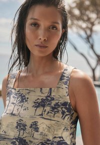 Top sans manches beige avec un col carré orné d'un motif paysager imprimé bleu. Le tissu semble léger et doux.