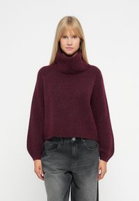 Maglione a collo alto borgogna con una vestibilità ampia, texture a costine e maniche larghe, abbinato a jeans grigi a vita alta.