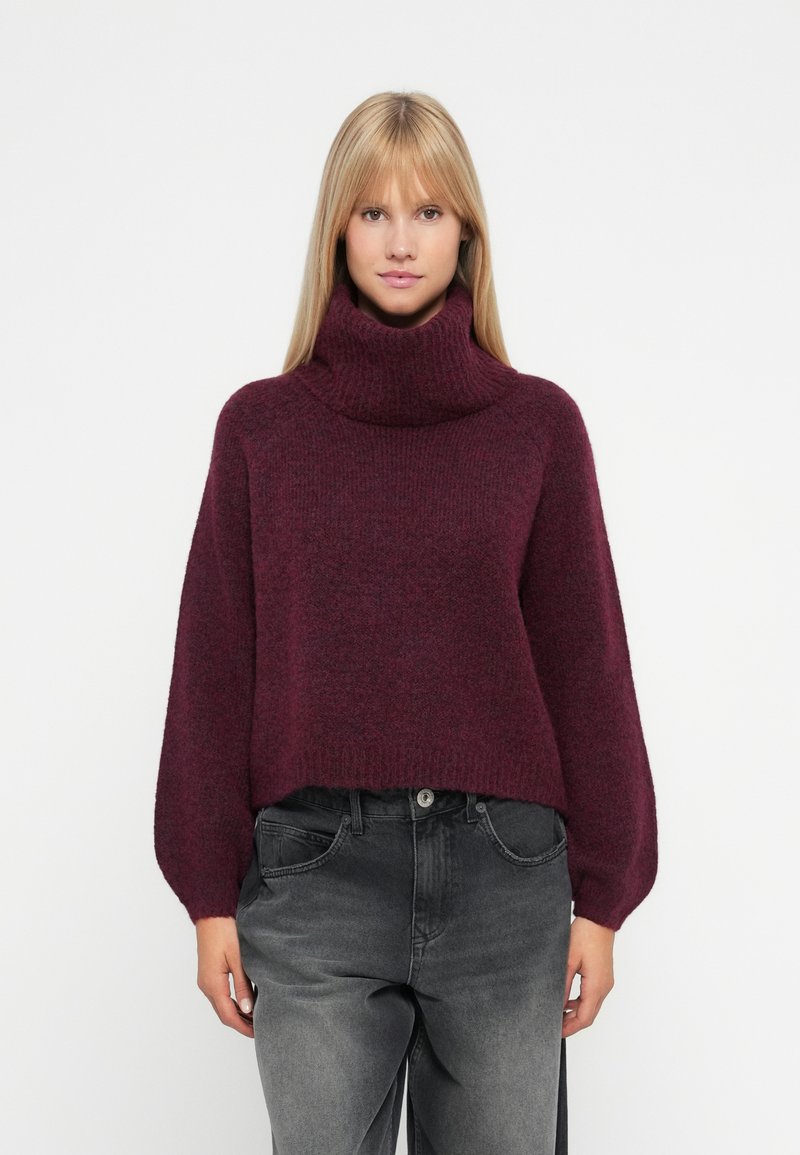 Maglione a collo alto borgogna con una vestibilità ampia, texture a costine e maniche larghe, abbinato a jeans grigi a vita alta.