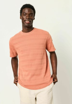 Smiling man wearing a textured coral short-sleeve shirt and light-colored pants, standing with hands in pockets against plain background.