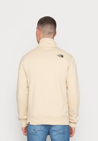 Pullover in pile beige con colletto alto, con logo nero sul retro. Polsini e vita a coste, texture liscia.