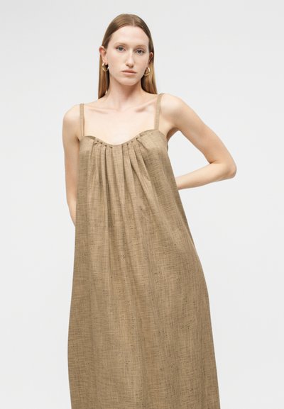 Γυναίκα με ίσια ανοιχτό καστανά μαλλιά που φοράει χρυσούς κρίκους στα αυτιά και μια χαλαρή, υφασμάτινη αμάνικη μπεζ φούστα με λεπτές τιράντες.