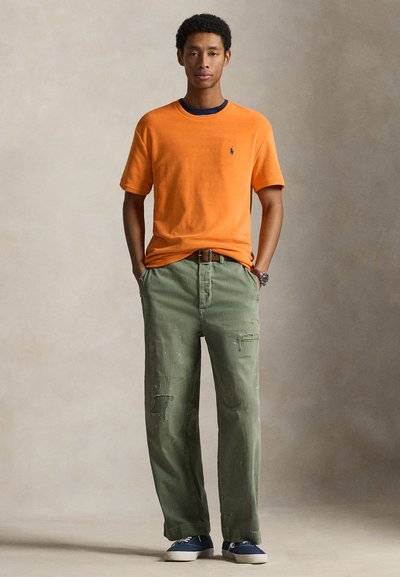 Άνδρας όρθιος με τα χέρια στις τσέπες, φοράει πορτοκαλί κοντομάνικο πουκάμισο, χακί σκισμένο παντελόνι, μπλε αθλητικά παπούτσια και ρολόι.