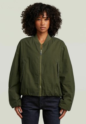 Frau mit lockigem Haar, die eine grüne Bomberjacke mit Reißverschluss und dunkle Hose trägt, steht vor einem hellen neutralen Hintergrund.