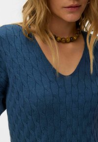 Plavi pleteni džemper s teksturiranim kabelskim uzorkom, V-izrezom i vidljivim šavovima. Okruglica od drvenih perlica upotpunjuje izgled.