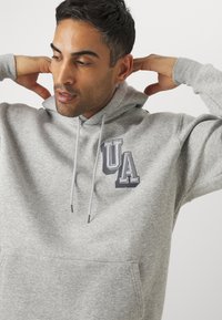 Sudadera gris hecha de una tela suave con bolsillo frontal, que presenta un logo "UA" bicolor en el pecho. Cuerdas de ajuste con extremos plateados.