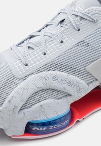 Vaaleanharmaa tennari, jossa on verkkomainen päällinen, teksturoidut yksityiskohdat, näkyvä "superrep"-brändäys ja sininen ilmatyyny punaisessa pohjassa.