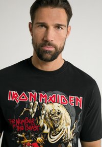 Černé bavlněné tričko s logem "Iron Maiden" a grafickým uměním kosterní postavy, s červenými a žlutými akcenty. Design s krátkými rukávy.