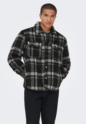 Uomo che indossa una giacca a quadri nera e grigia con tasche sul petto, in piedi con le mani nelle tasche laterali contro uno sfondo semplice.