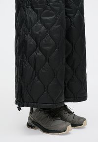 Juoda siuvinėta kelnė su blizgia tekstūra, su susiaurintu apačiu. Derinama su tamsiai pilkais žygio bateliais, turinčiais tekstūruotą tinklinę medžiagą ir guminiu padu.