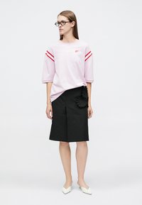 Γυναίκα που φοράει ροζ μπλούζα με μανίκια μέχρι τον αγκώνα και κόκκινες ρίγες, μαύρο σορτς μέχρι το γόνατο με μαύρα λουλουδένια σχέδια, άσπρα μυτερά παπούτσια και γυαλιά.