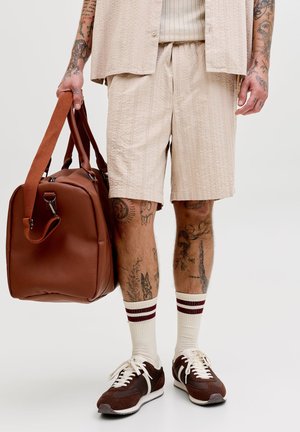 Férfi bézs rövidnadrágban és ingben, fehér zokniban barna csíkokkal, barna sportcipőben, kezében egy nagy barna bőr utazótáskával, karjain és lábain látható tetoválásokkal.