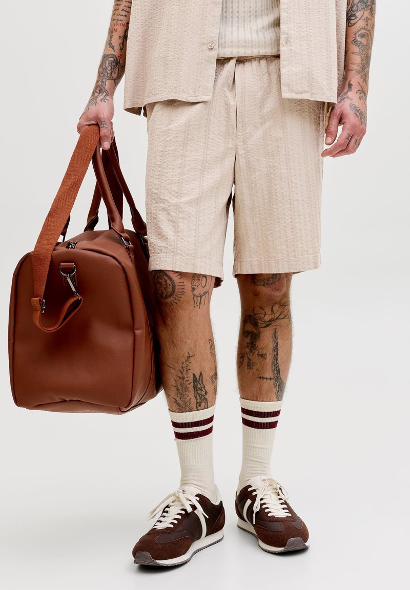 Muškarac u bež kratkim hlačama i majici, bijele čarape s smeđim prugama, smeđim tenisicama, drži veliki smeđi kožni sportski torbu, vidljive tetovaže na rukama i nogama.