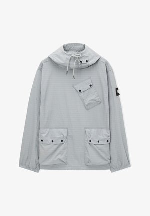 Vaaleanharmaa, kevyt takki, jossa on korkea kaulus ja kiristysnyöri. Takissa on yksi vino rintatasku sekä kaksi etutaskua, joissa on mustat napit.