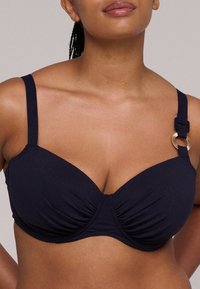 Marineblauwe bikini-top met een ruchesdesign, beugelsteun, dunne bandjes en accenten van goudkleurige hardware. Glad textuur.