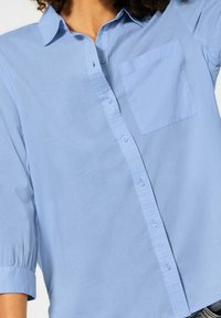 Světle modrá košile na knoflíčky s náprsní kapsou, krátkým rukávem s nařasenými manžetami a hladkou, jemnou texturou.