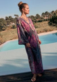 Rochie maxi vaporoasă cu model paisley în nuanțe de mov, roz și teal. Are mâneci clopot și decolteu în V. Asortată cu sandale aurii.