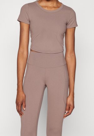 Femme portant un crop top taupe à manches courtes et un legging taille haute assorti, debout sur un fond blanc uni.