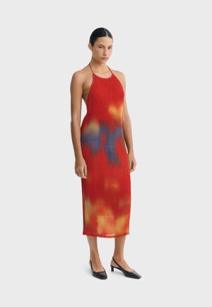 Femme aux cheveux foncés portant une robe midi sans manches rouge à col halter avec des motifs abstraits orange et bleus, chaussée de chaussures noires à petit talon.